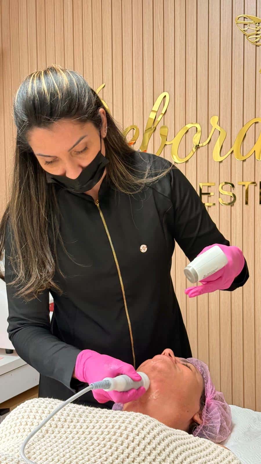 Debora, in a black uniform and pink gloves, gives a facial treatment to a client lying on the treatment bed, in front of the gold studio sign.